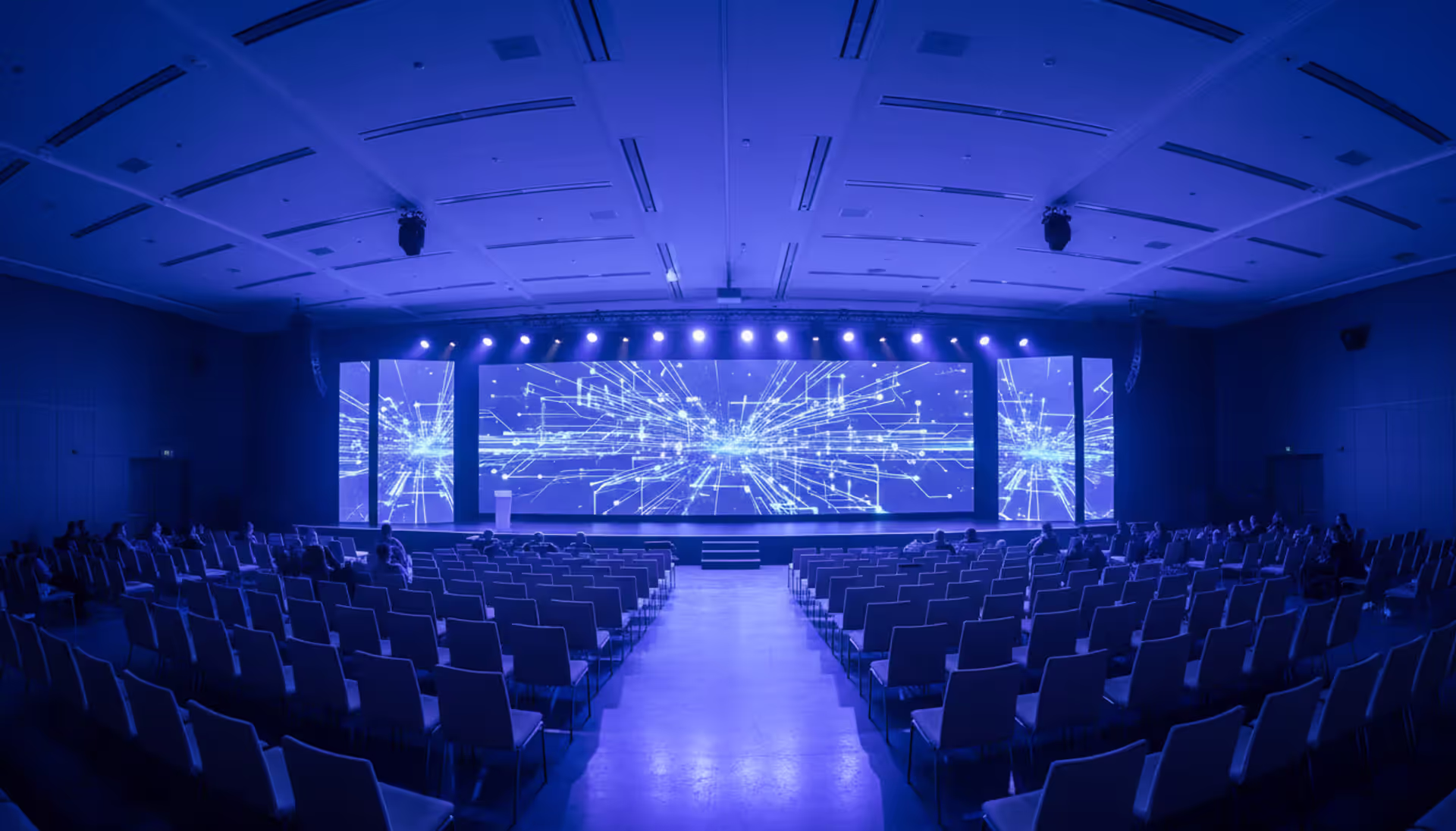 Modern conference hall with a large stage, blue and purple lighting, digital network graphics on the screen, and audience silhouettes
