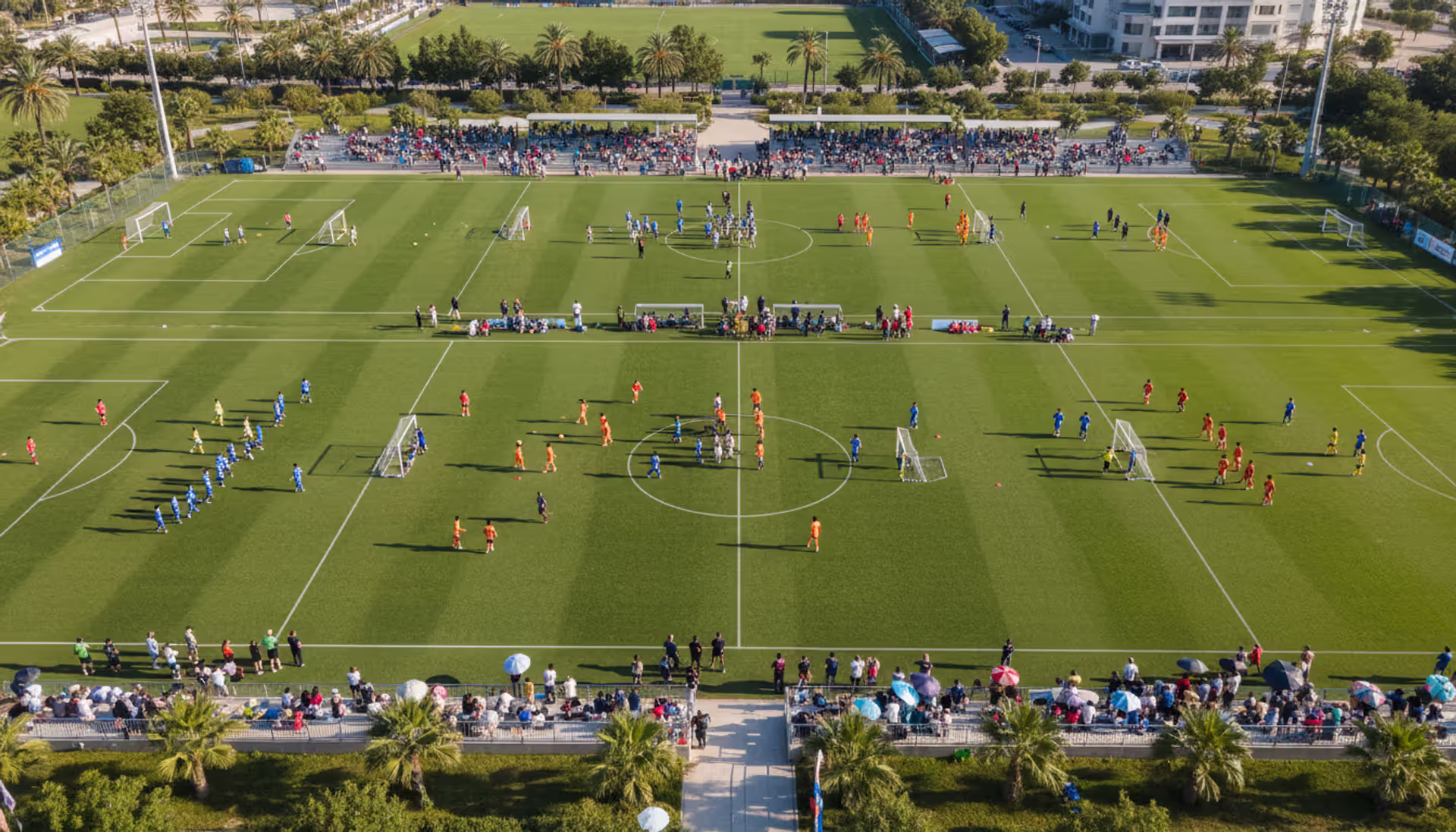 Aerial view of a large youth soccer tournament with multiple fields, teams in colorful uniforms playing simultaneous matches, spectators on sidelines on a sunny day